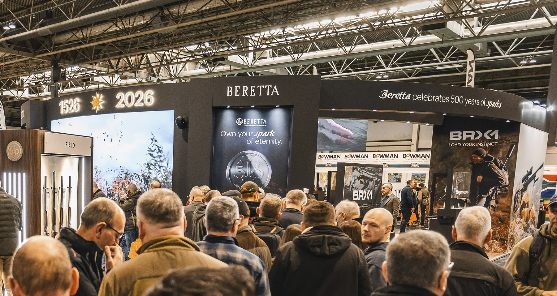 A GMK team member holding a camouflage-finish Beretta shotgun in front of the 1526 to 2026 anniversary screen
