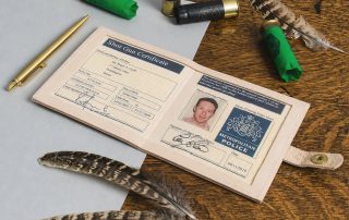 Shot gun certificate open in its leather wallet on a dark oak table, with three shotgun cartridges, a brass pen and two partridge feathers