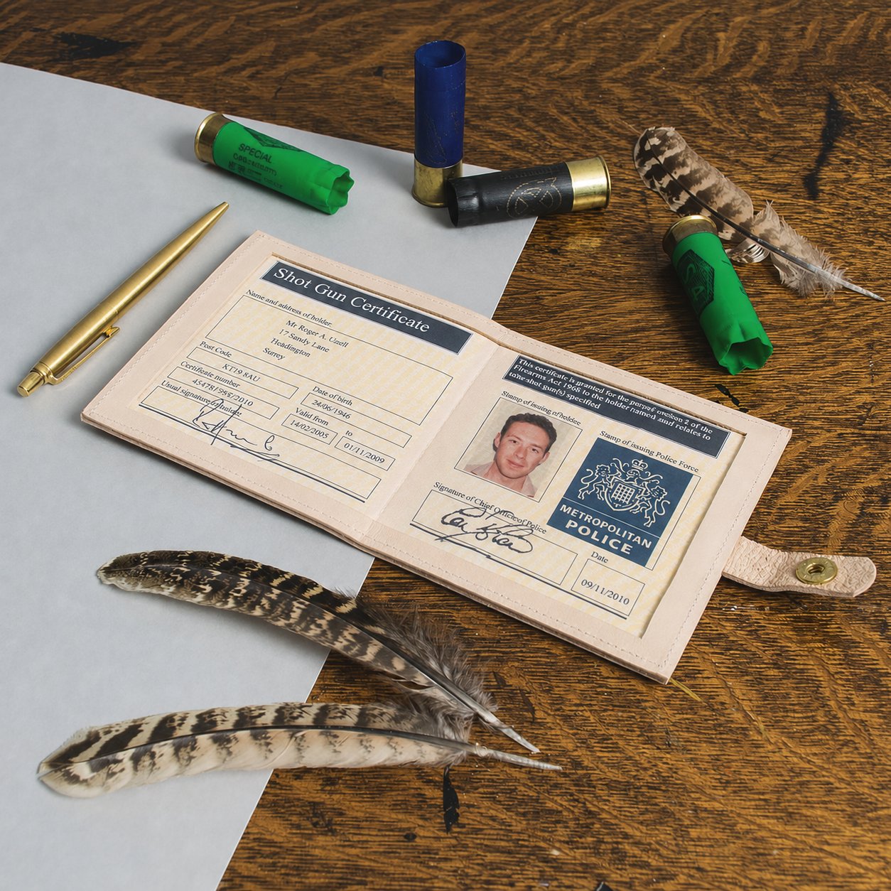 Shot gun certificate open in its leather wallet on a dark oak table, with three shotgun cartridges, a brass pen and two partridge feathers