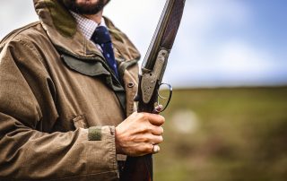 A Gun in a shooting jacket and tie holding a broken side-by-side shotgun on a hill