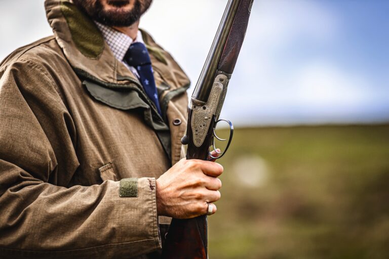 A Gun in a shooting jacket and tie holding a broken side-by-side shotgun on a hill