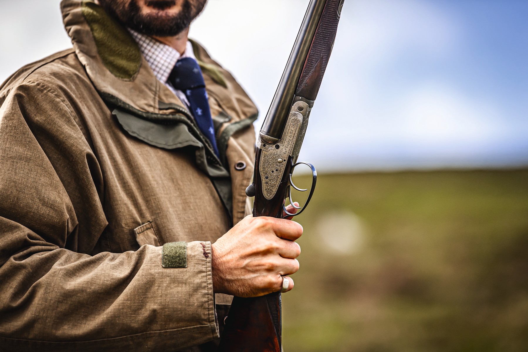 A Gun in a shooting jacket and tie holding a broken side-by-side shotgun on a hill