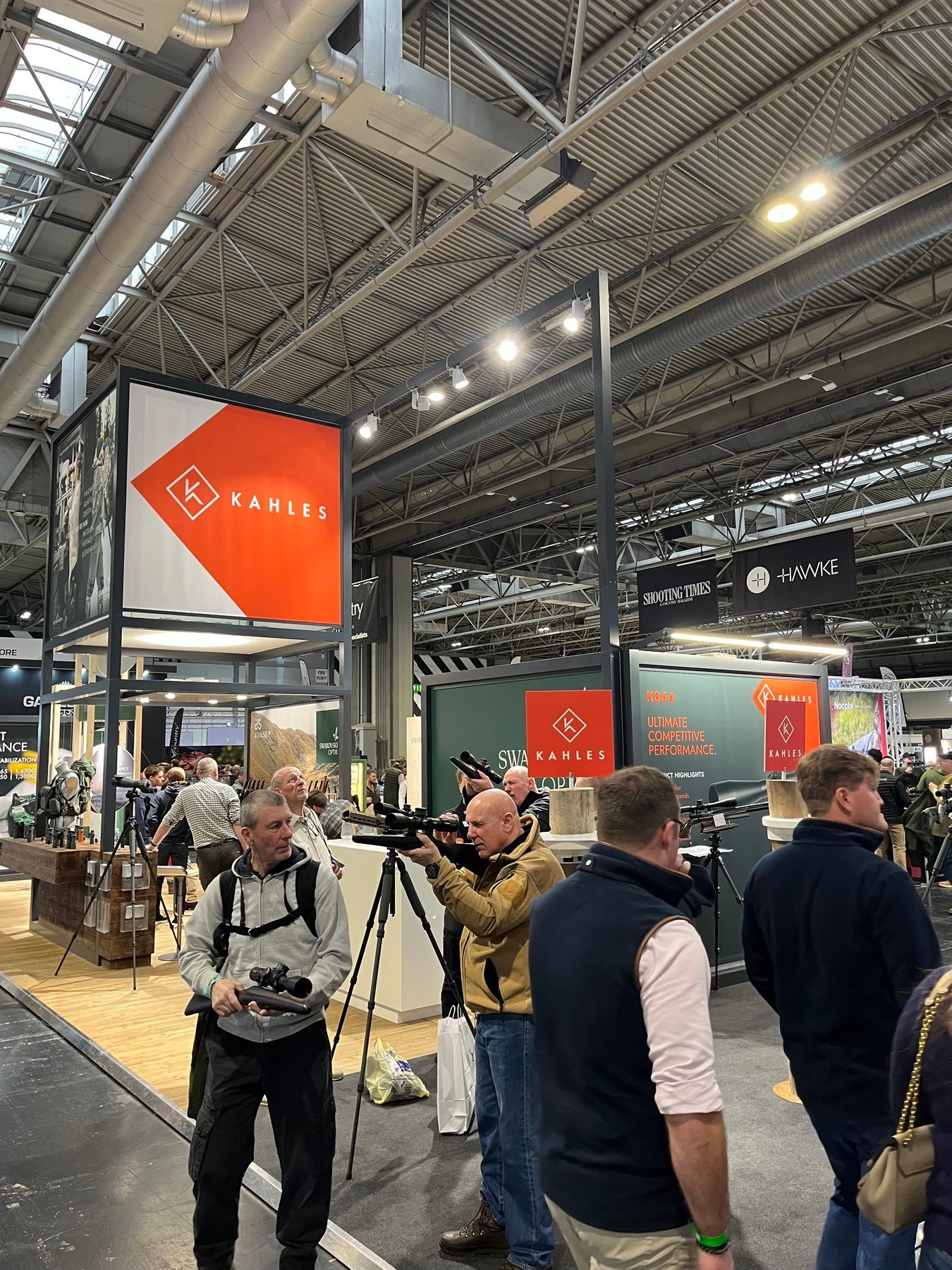 A visitor at a tripod-mounted spotting scope beneath the Kahles branding at the British Shooting Show